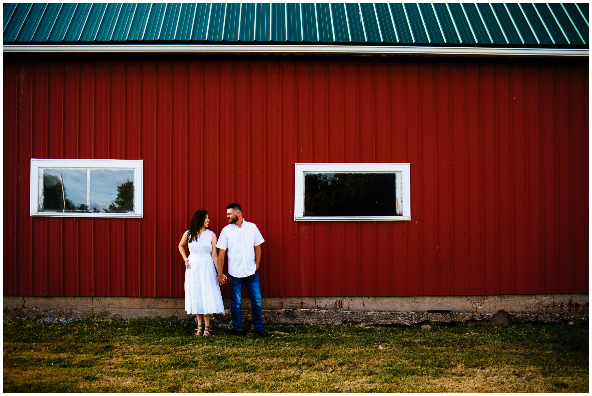 A woman wears a white dress to her farm engagement session. She looks at her fiancé and smiles. They are in front of a red barn.
