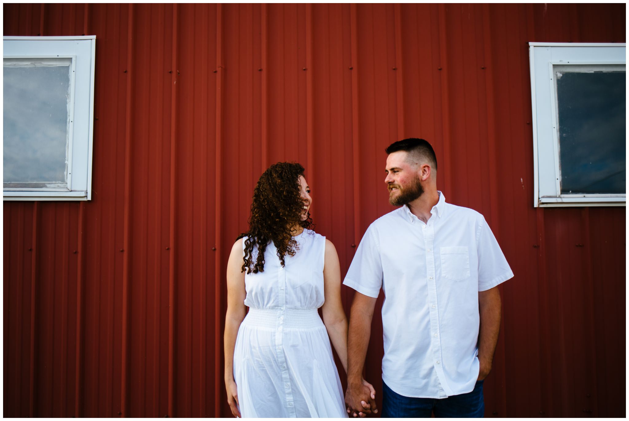A man and woman smile at each other in front of a red barn, during their summer engagement session.