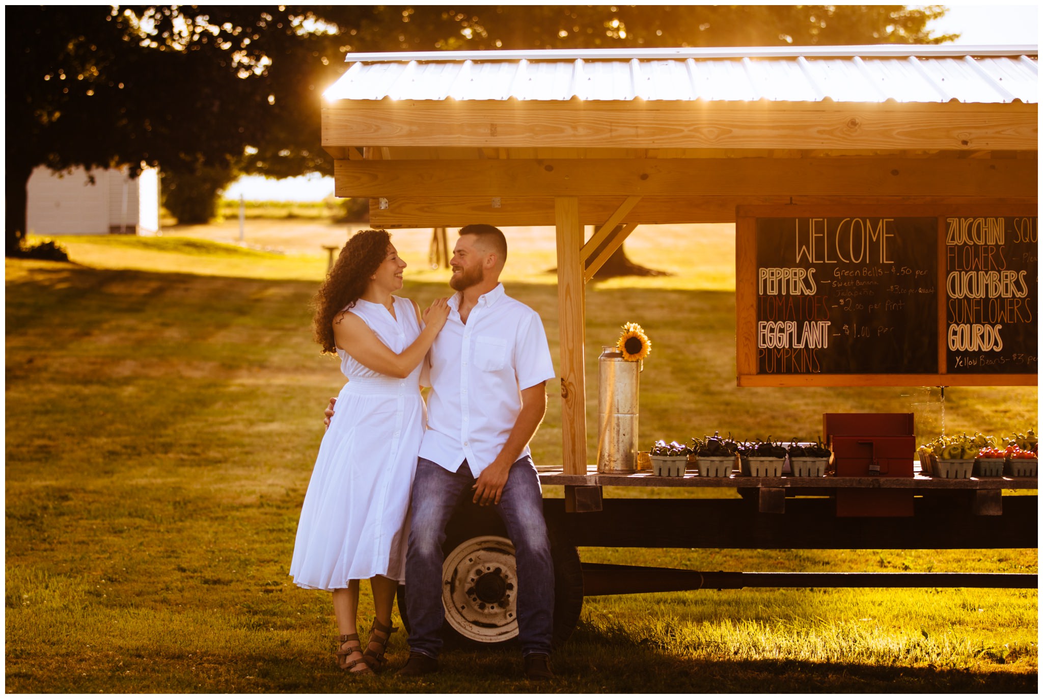Golden sunlight falls during a flower farm engagement session. The bride to be and groom to be sit on a farm stand that is full of produce.