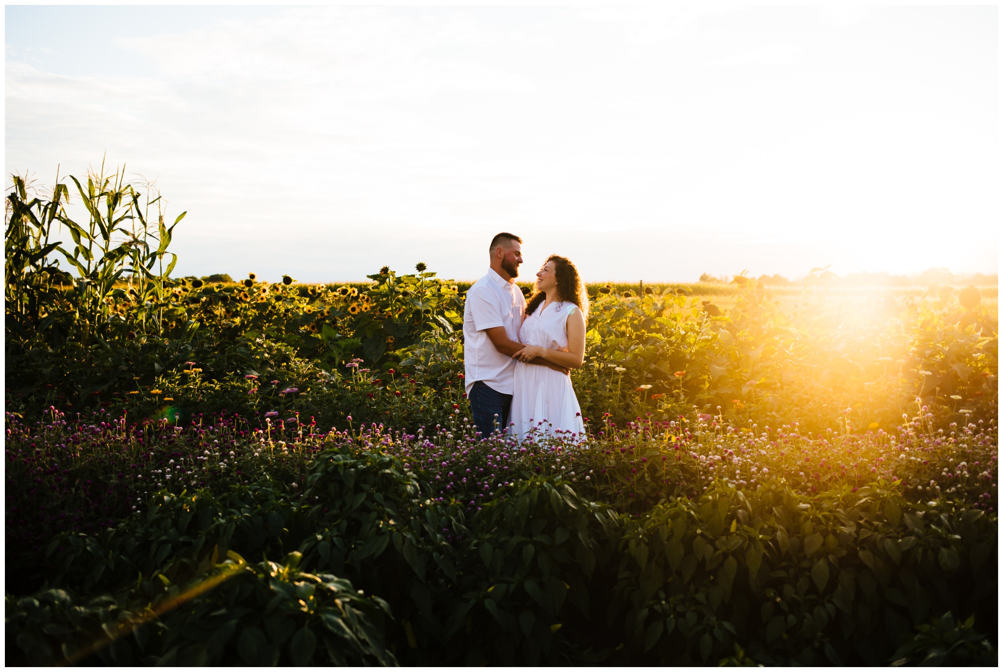 A couple gazes at each other in a field of wildflowers during their summer engagement session on a farm.