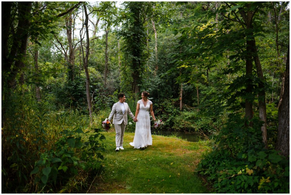 The brides walk through the woods near a stream, holding hands at their Sinclair of Skaneateles wedding.