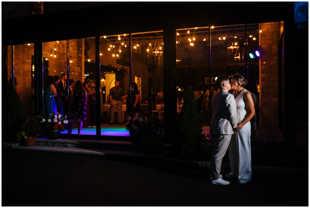 The brides stand outside the Sinclair of Skaneateles at night, sharing a kiss. Inside, guests dance on the dance floor. 