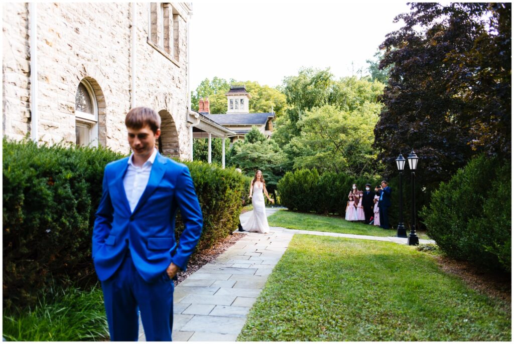 Groom in a blue suit waits as his bride walks up to him from behind for their first look on their wedding day. The first look took place outside at Fountain Place Ithaca.