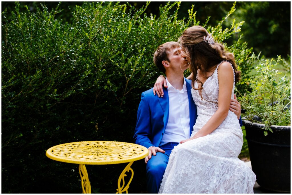 Bride and groom sit at a bright yellow table outside and share a kiss on their wedding day.