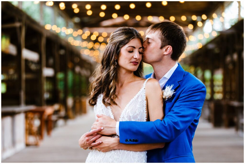 Beautiful image of groom kissing bride on the cheek at their Ithaca Farmer's Market wedding. Twinkle lights create bokeh behind them.