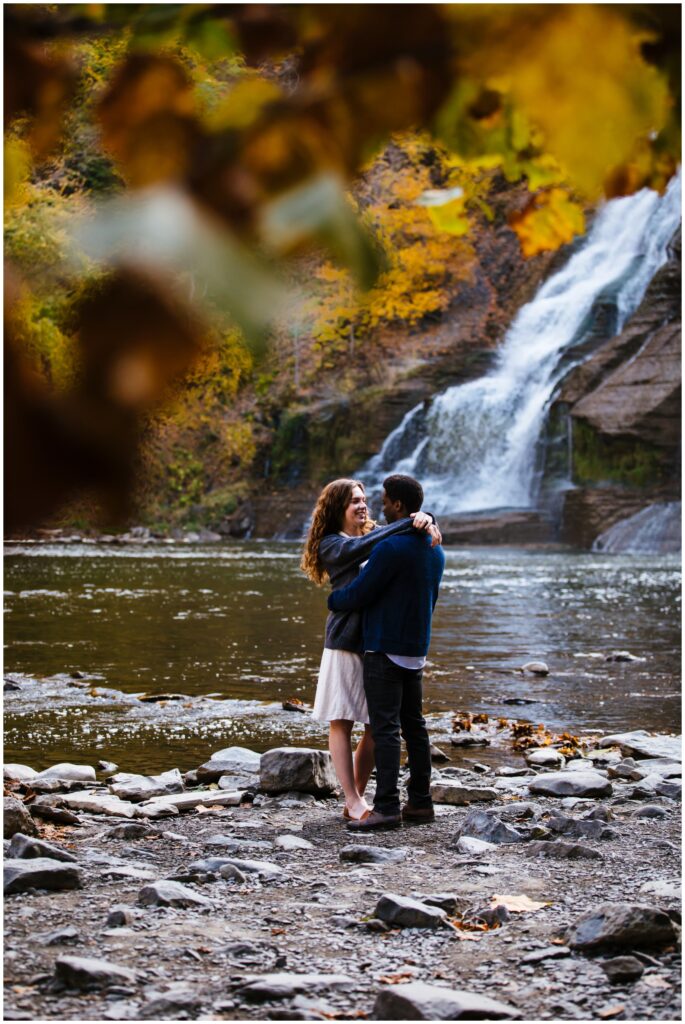 Couple looks at each other during their ithaca engagement session with a waterfall in the background.