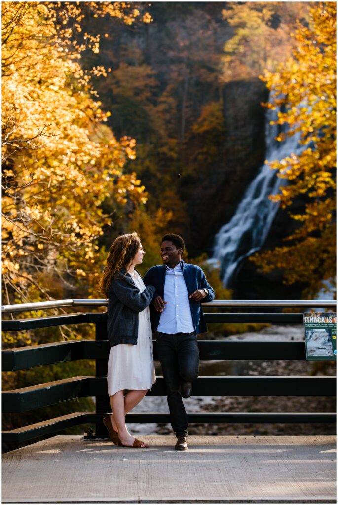 Couple stands on a bridge looking at each other during their ithaca engagement session. The waterfall is in the background.
