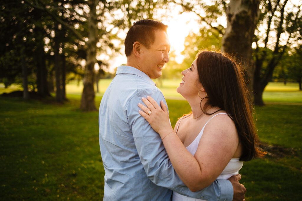 A couple smiles at each other as the sun streams in from behind during their rochester new york engagement session.