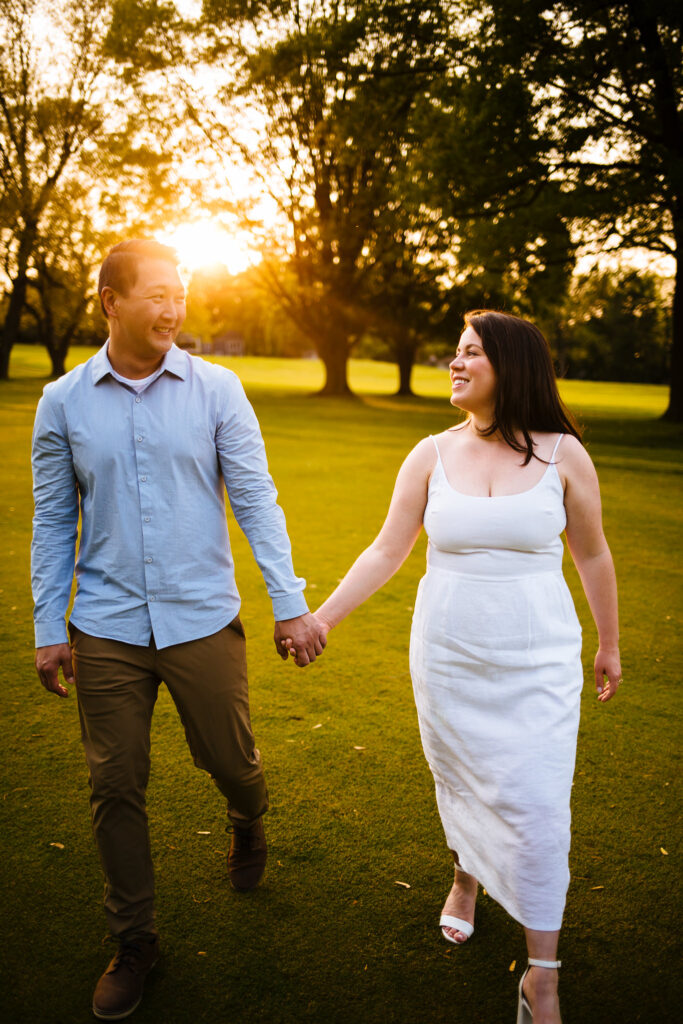 couple holds hands and walks towards the camera, the sun is setting behind them.