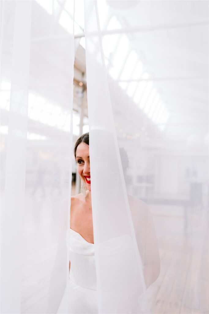 Bride peeks at groom through sheer curtain.