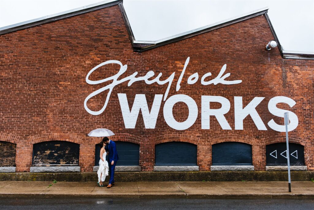 Bride and groom stand under an umbrella and kiss with Greylock works sign behind them.