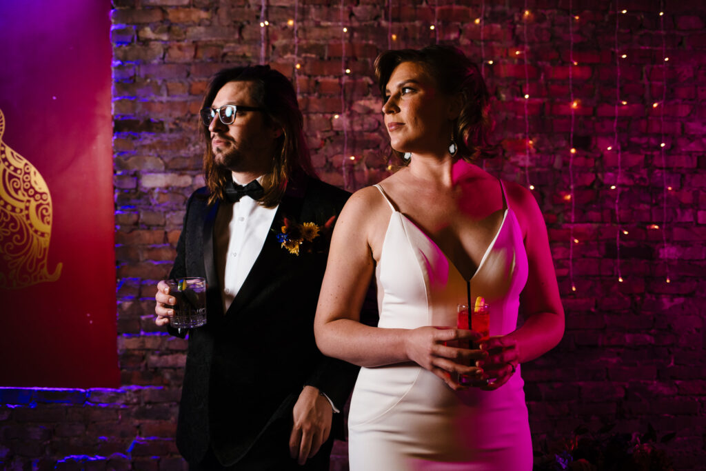 Bride and groom stand in front of a brick wall with string lights. They look off to the side. They are having their wedding at the Gilded club in Syracuse, NY which has a speakeasy vibe.