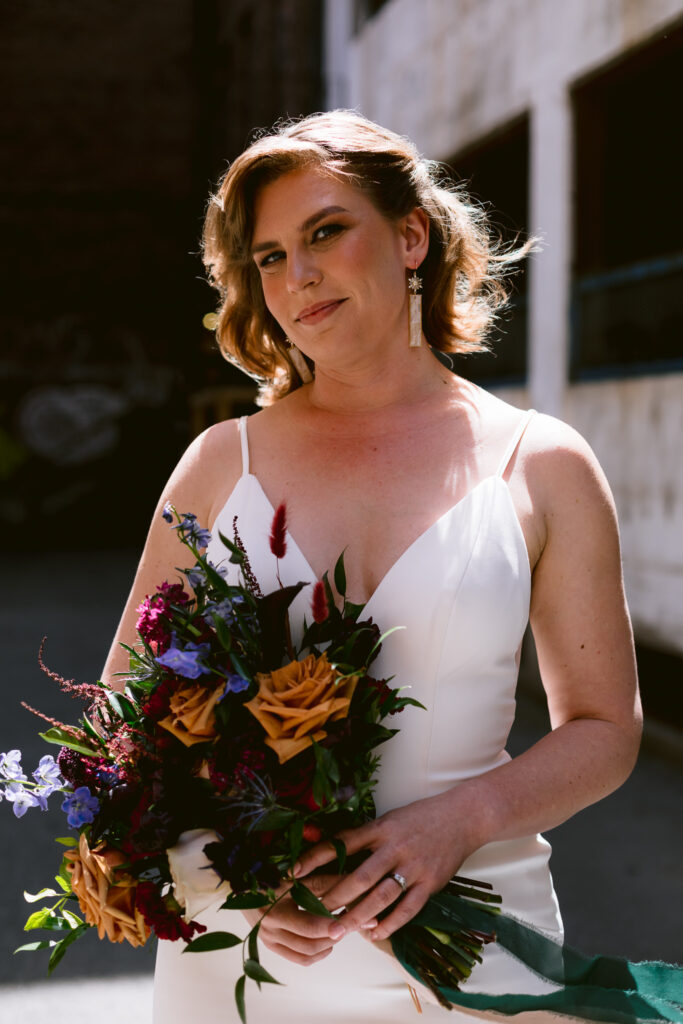 Bride stares at camera. She is outside and holding a colorful jewel toned bouquet. She is next to an industrial building.