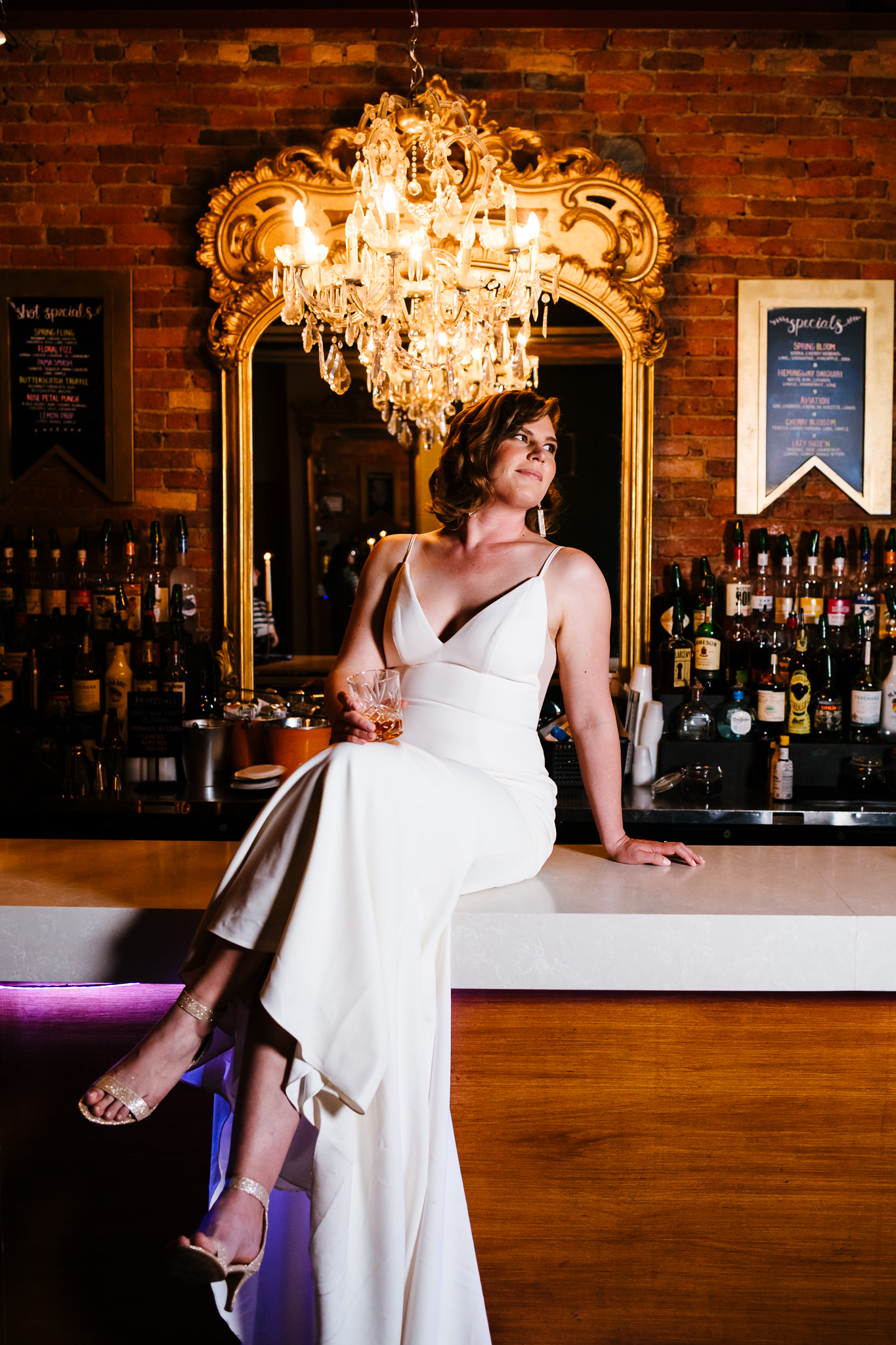 Bride sits on the bar of the Gilded club in Syracuse, NY. She is holding a drink and looking off camera.