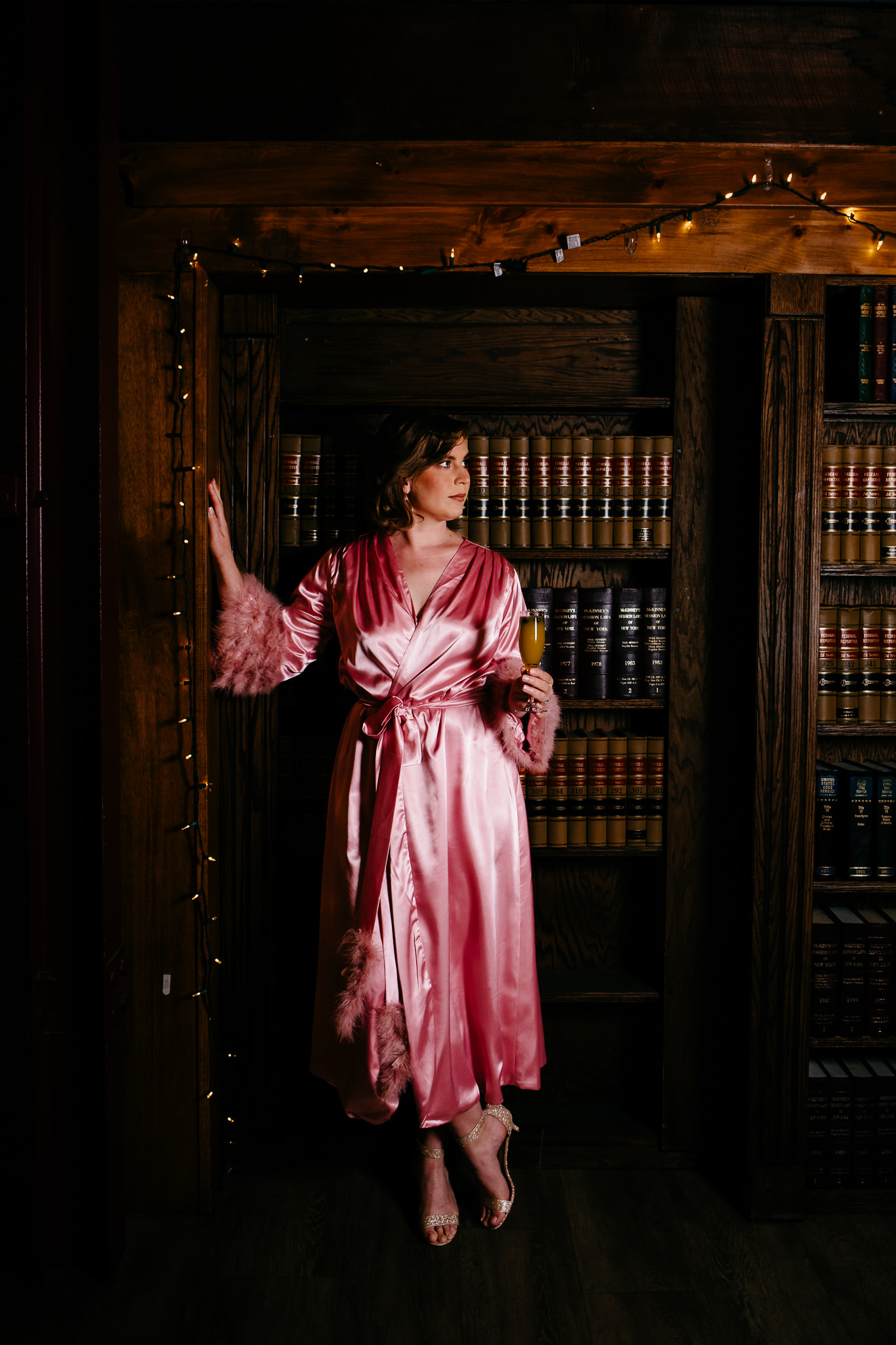 Woman wears a pink robe while getting ready at the Gilded Club in Syracuse, NY. She sips on a mimosa and leans against a shelf.