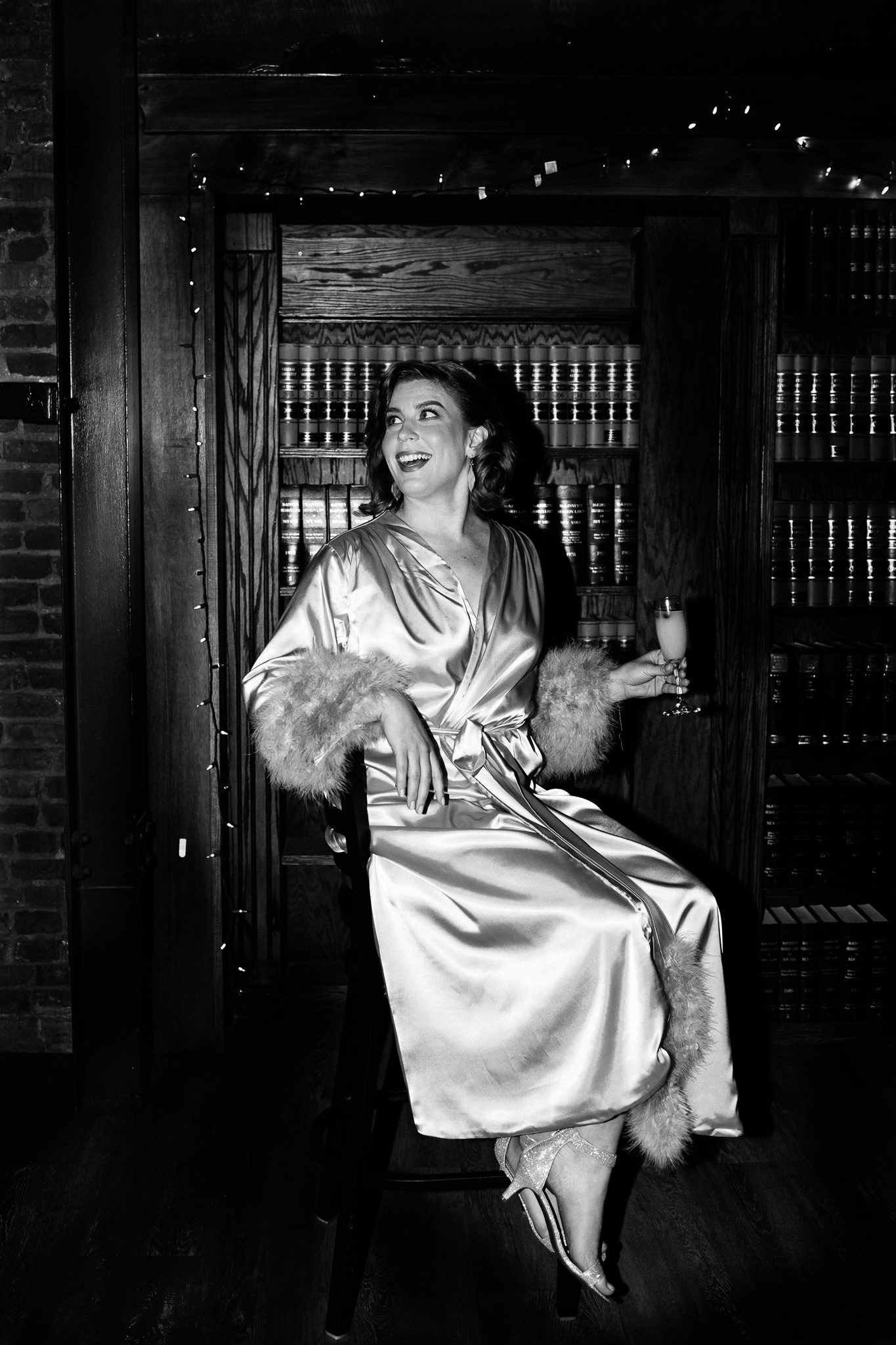 Black and white photo of bride in a robe holding a mimosa. A candid wedding photo at the Syracuse Gilded Club. She is laughing.