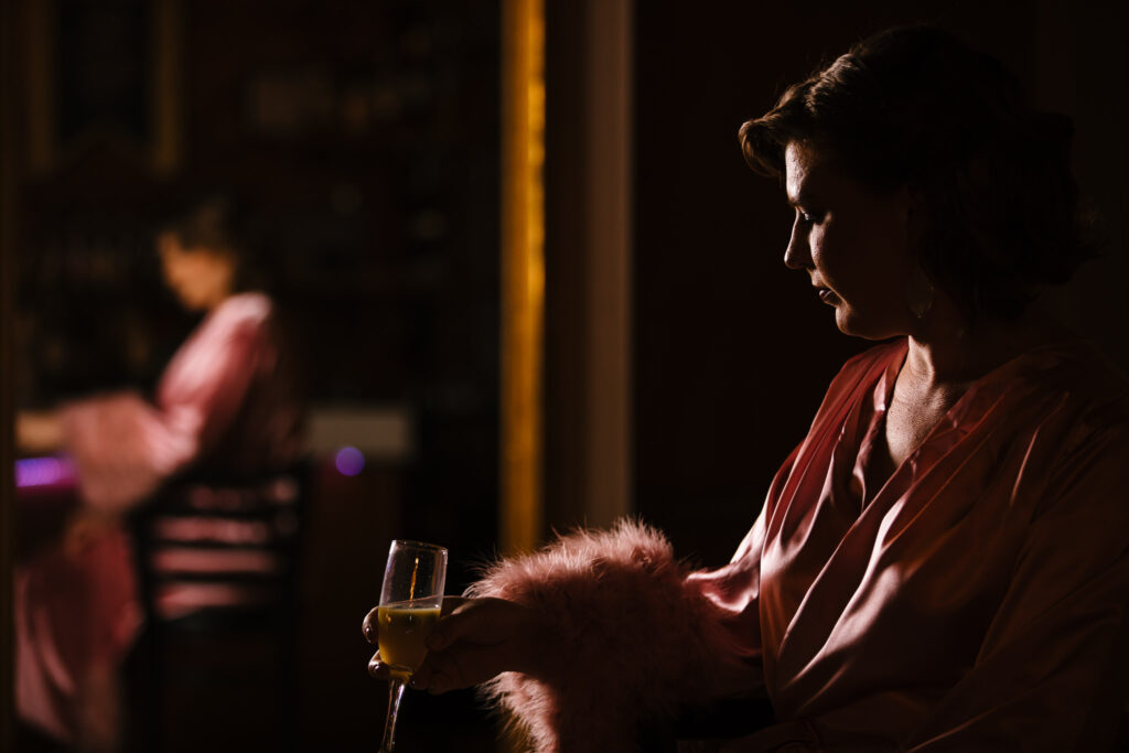 Woman wears a pink robe while getting ready at the Gilded Club in Syracuse, NY. She holds a mimosa.