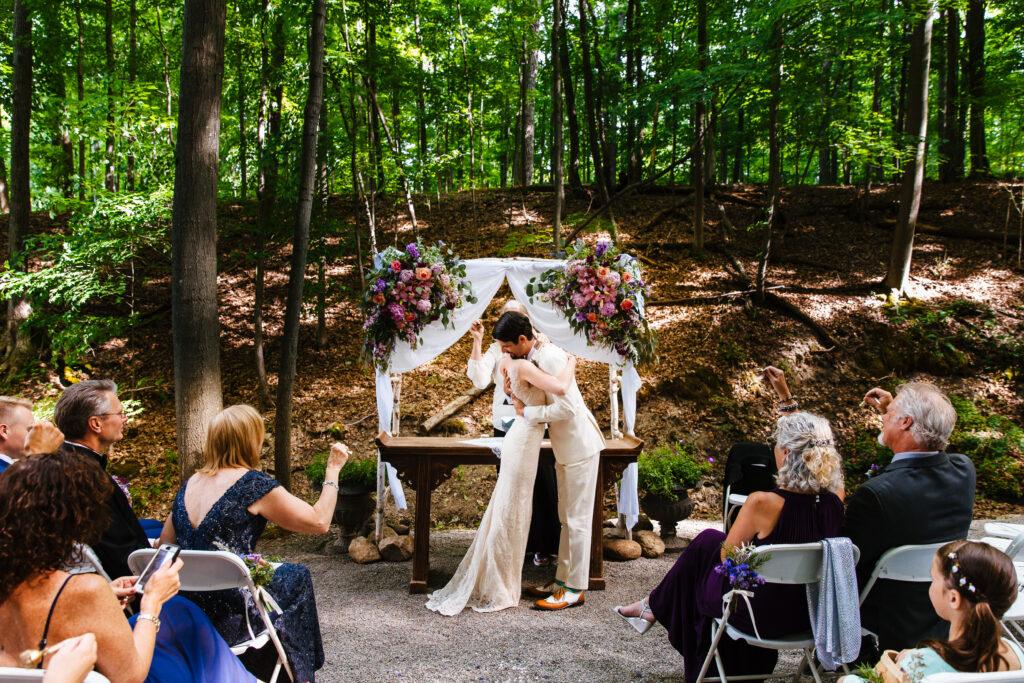 Bride and groom embrace during their wedding ceremony in the woods. Behind them their wedding Arbor is surrounded by flowers.