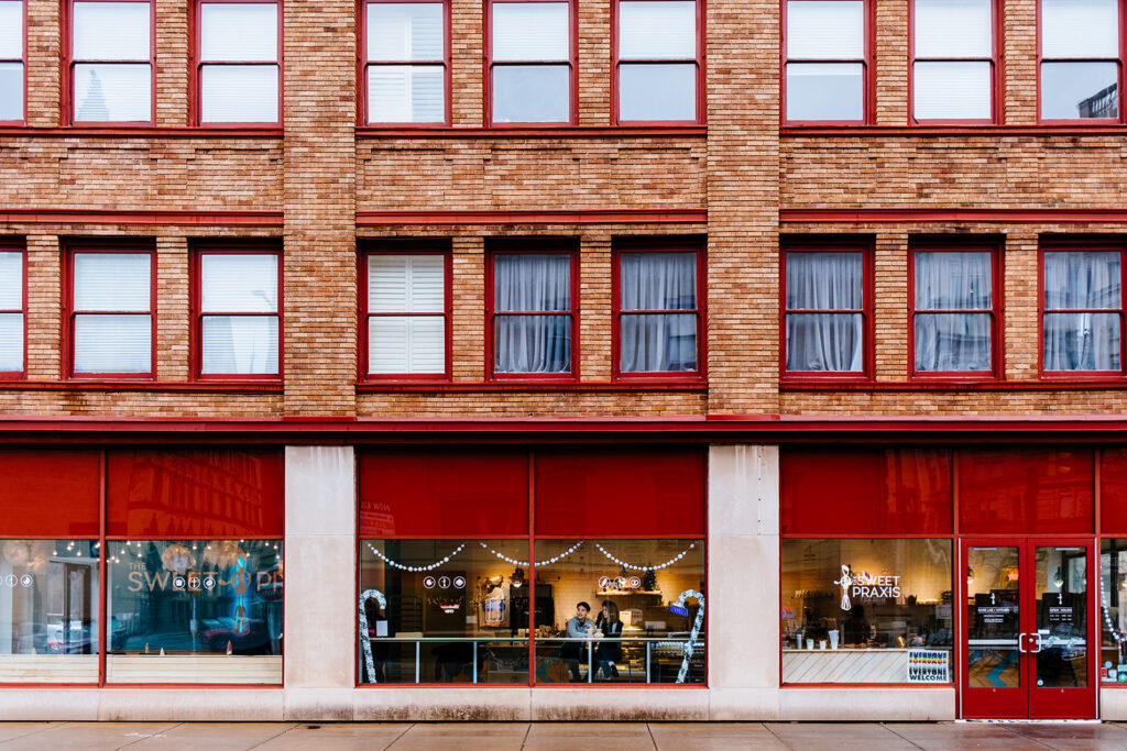 An image of the couple through a coffee shop window. They are at Sweet Praxis in Syracuse, NY.  They are smiling. 