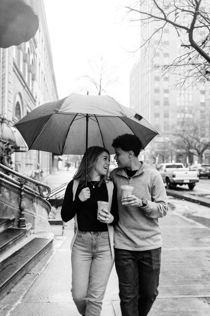 Bride and Bride carry coffee and walk down the street under an umbrella .