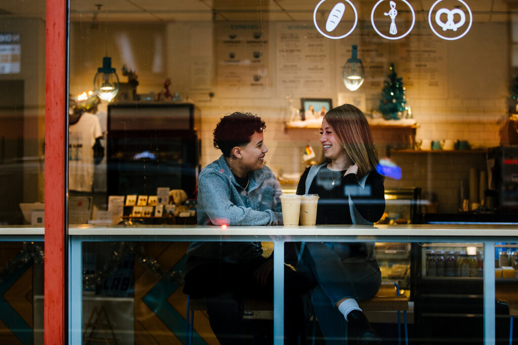 An image of the couple through a coffee shop window. They are at Sweet Praxis in downtown Syracuse, NY. This is during their winter Syracuse Engagement Session. They are smiling. This is a LBGTQ engagement session.