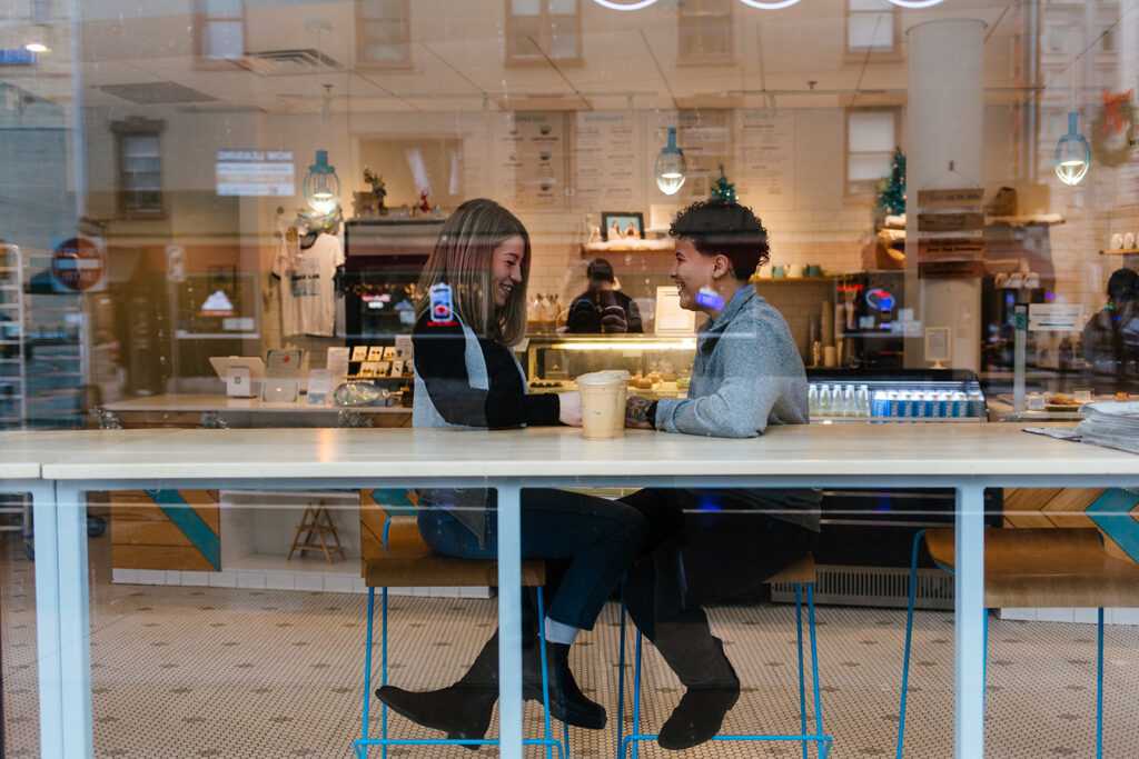 An image of the couple through a coffee shop window. They are at Sweet Praxis in downtown Syracuse, NY. This is during their winter Syracuse Engagement Session. They are smiling. This is a LBGTQ engagement session.