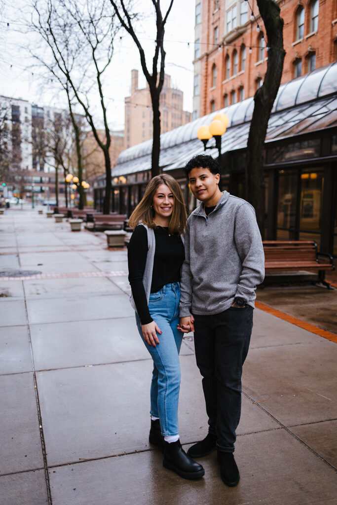 Bride and bride hold hands and smile at each other during their winter engagement session in downtown syracuse.