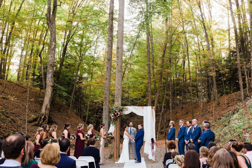 The bride and groom stand at a floral arbor in the woods at New Park Event Venue