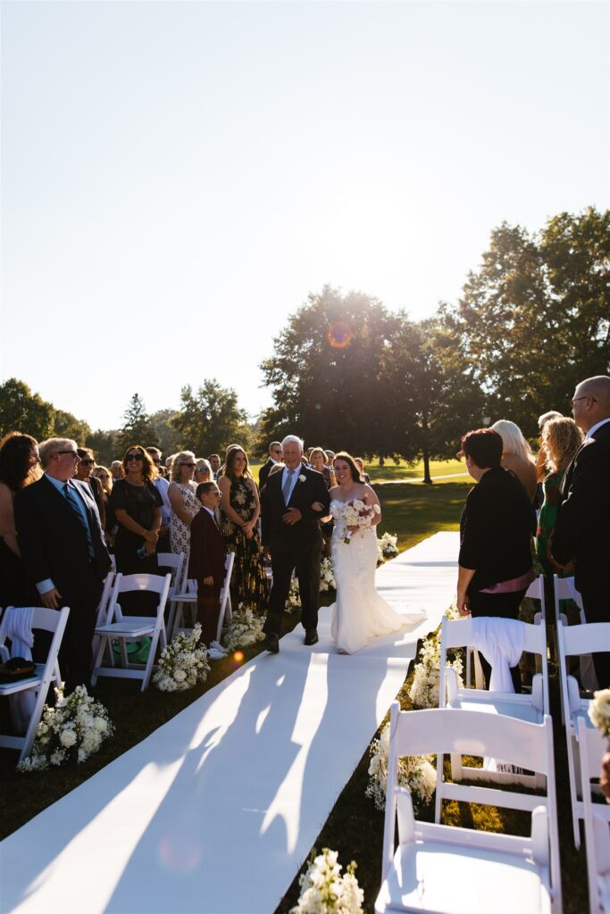 A bride walks down the aisle with her father as golden light streams behind them. Her wedding is in late September in Central NY. Photographed by Central NY wedding photographer, Calypso Rae Photography.