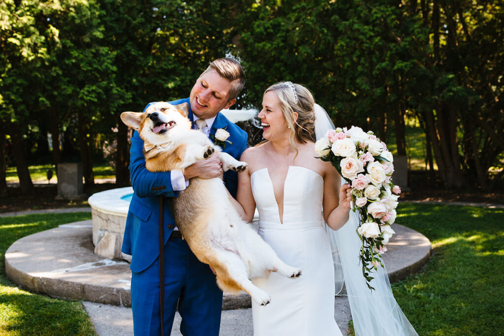 A newlywed couple smiling at a dog in one of their arms 