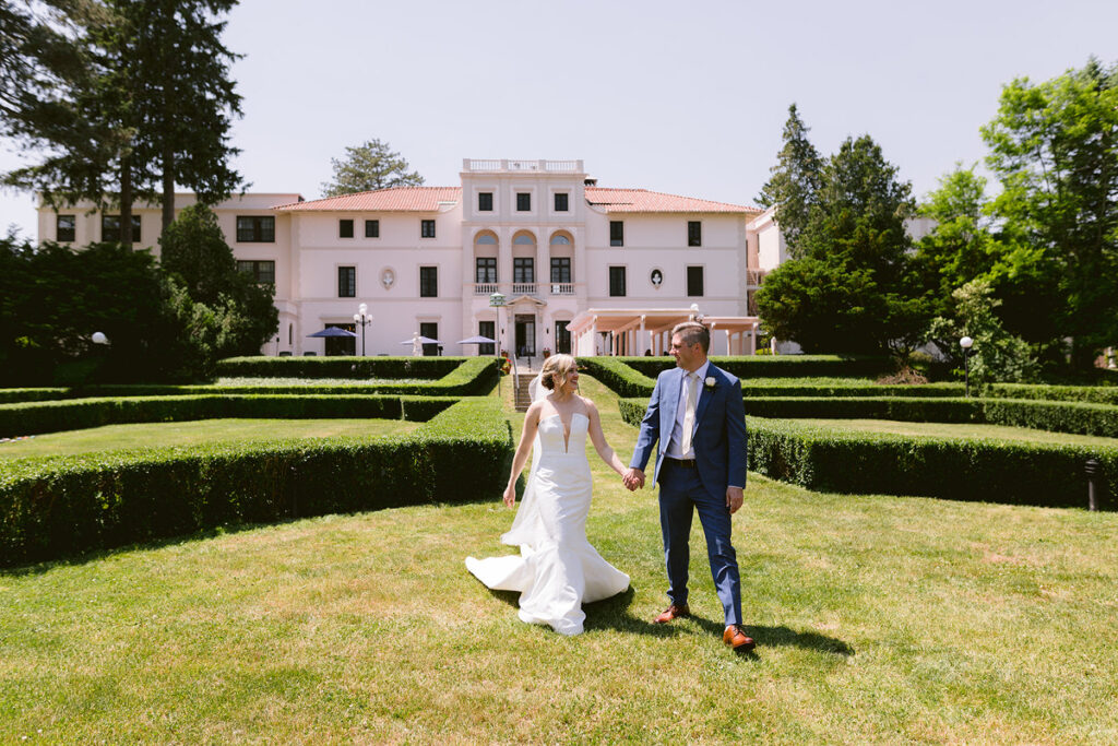 A newlywed couple holding hands and walking in a garden with a mansion behind them 