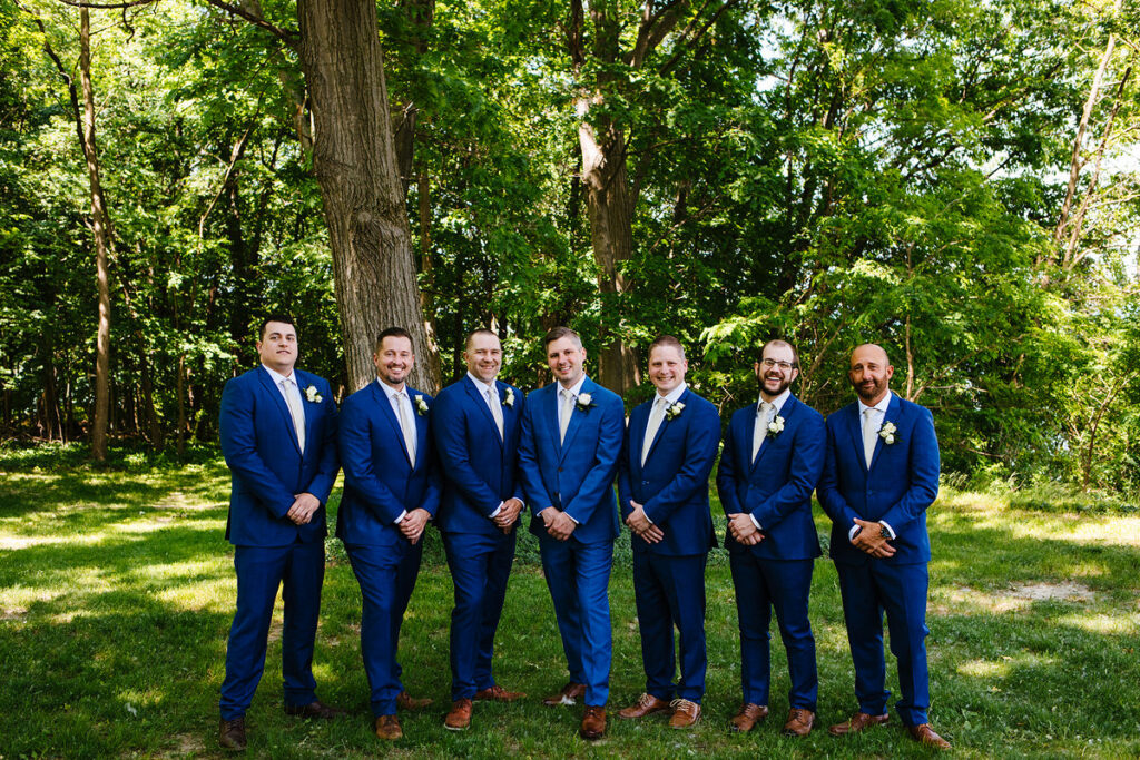 A wedding party in matching blue suits smiling in a wooded area 