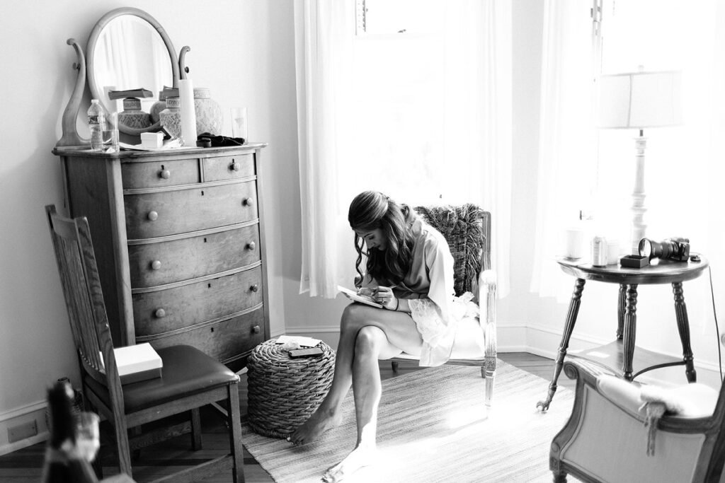 A black-and-white image of a bride sitting on a chair in a bright room, writing a letter, with a wooden dresser and a round mirror behind her.