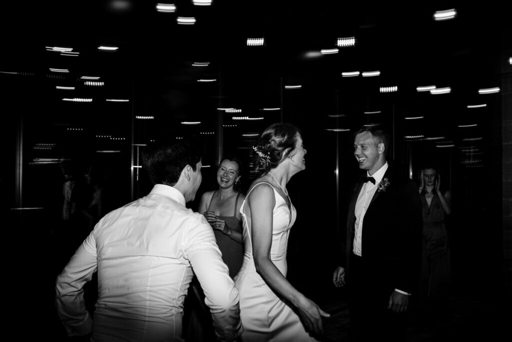 A black-and-white photo of guests dancing at a wedding reception, with the bride and groom smiling at each other.