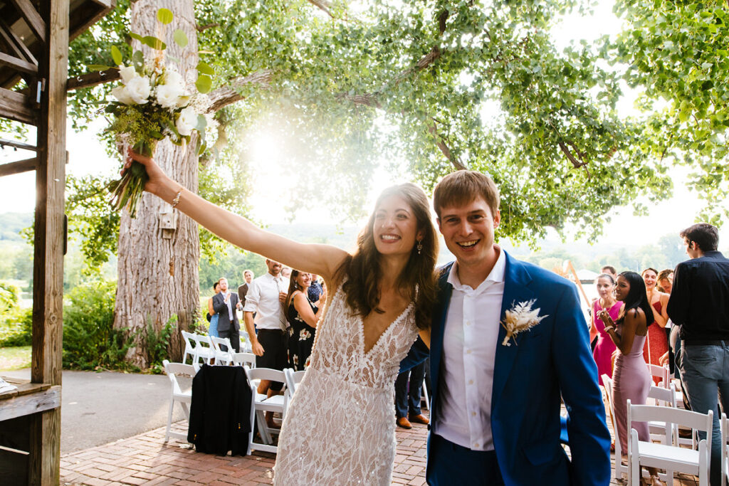 A bride and groom smiling as they walk together, with the bride holding a bouquet up and guests in the background.