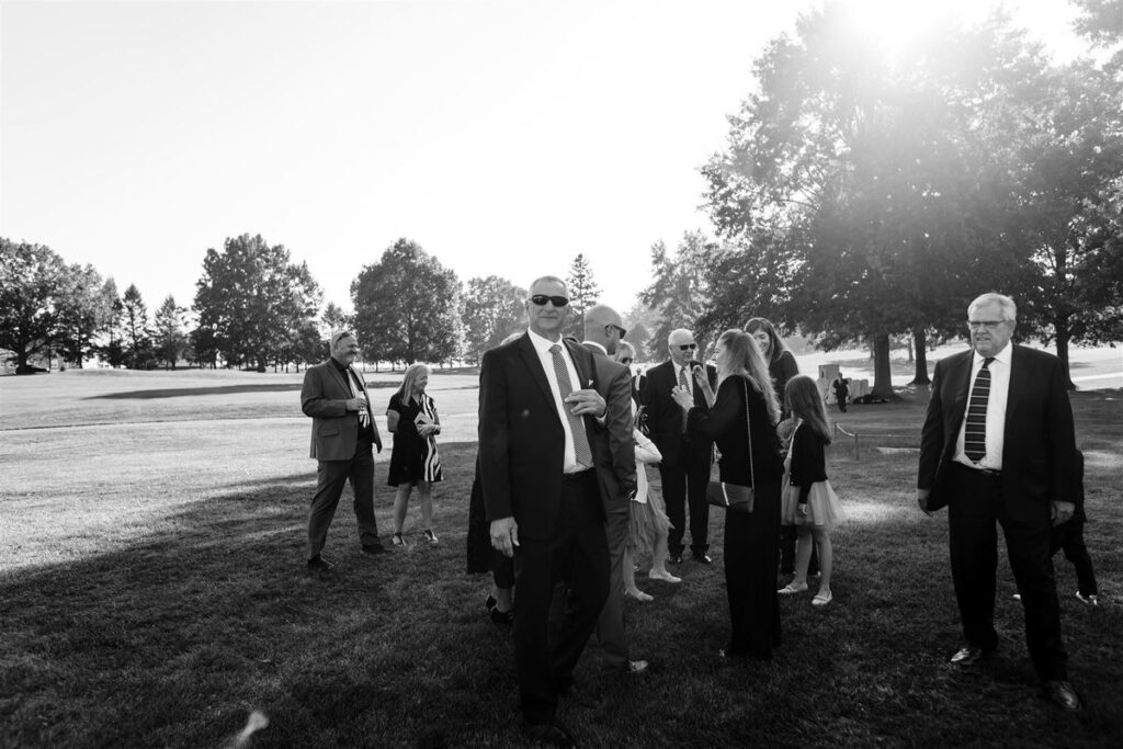 A group of wedding guests standing on a lawn with trees in the background.