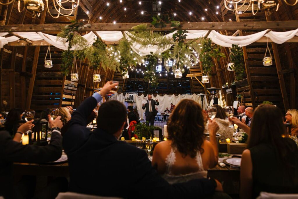 Guests raising glasses in a toast during a wedding reception in a barn with string lights and greenery.