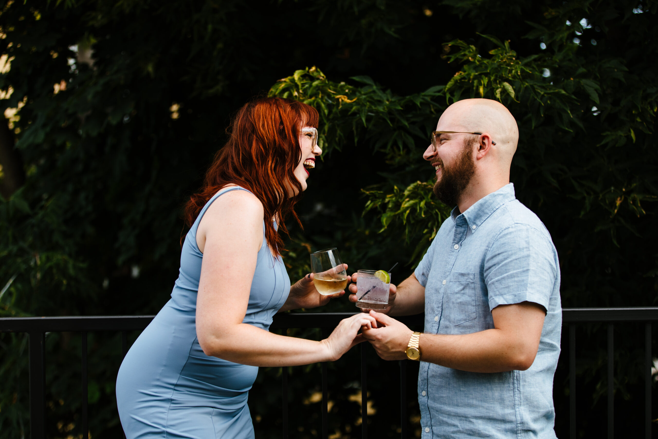 A woman in a blue dress with red hair and glasses is holding a drink in her hand and holding the hand of a man and laughing while the man in a blue shirt is also holding a drink and smiling and laughing with the woman