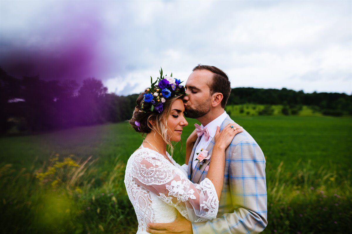 Colorful wedding photography showing a person kissing their partner's head with purple smoke next to them.