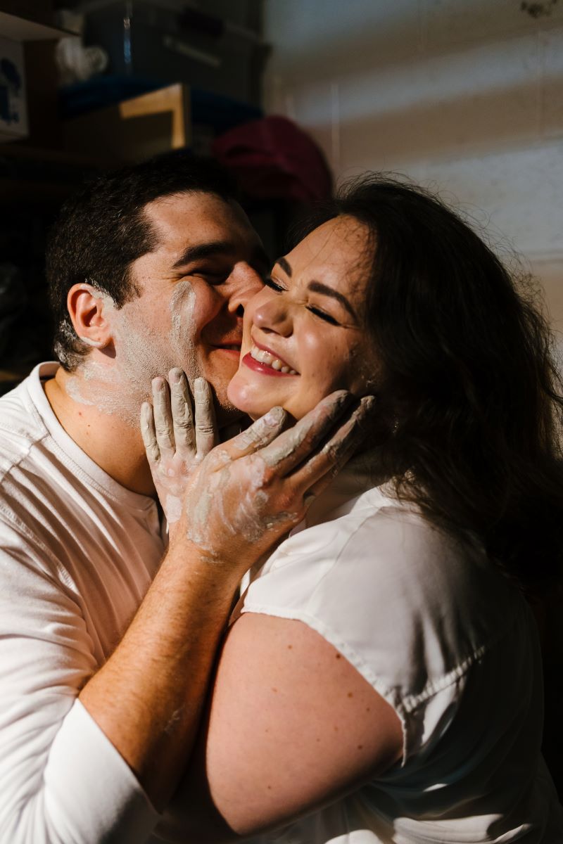 A man with pottery clay on his hands giving a woman a kiss on the cheek the woman is smiling and she also has pottery clay on her hands and is touching the face on the man both are wearing white shirts 