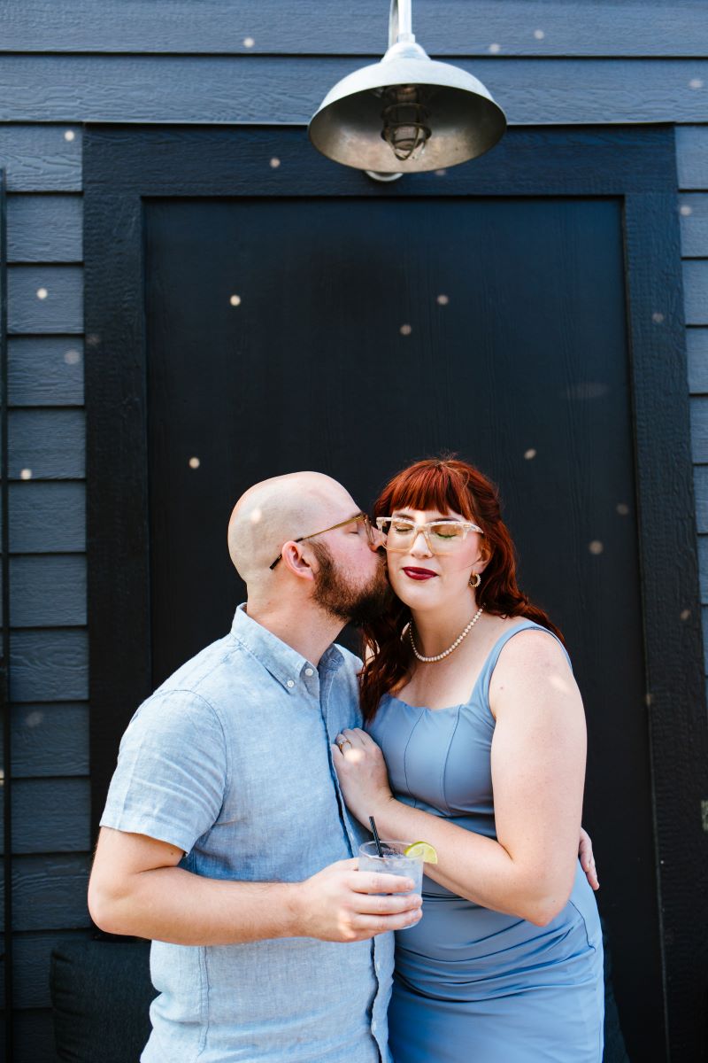 A man in a blue shirt holding a drink  with a lime in his hand giving a woman with red hair a glasses and a blue dress a kiss on the cheek 