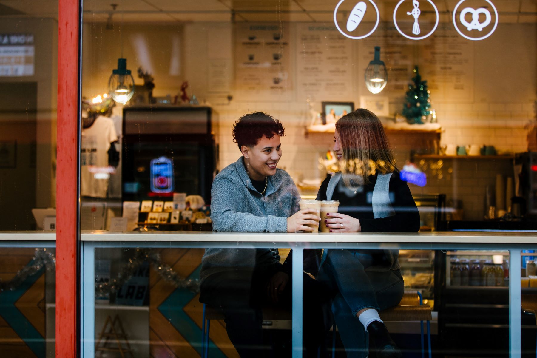 A couple sitting in a coffee shp holding coffees smiling and laughing behind a glass window pane 