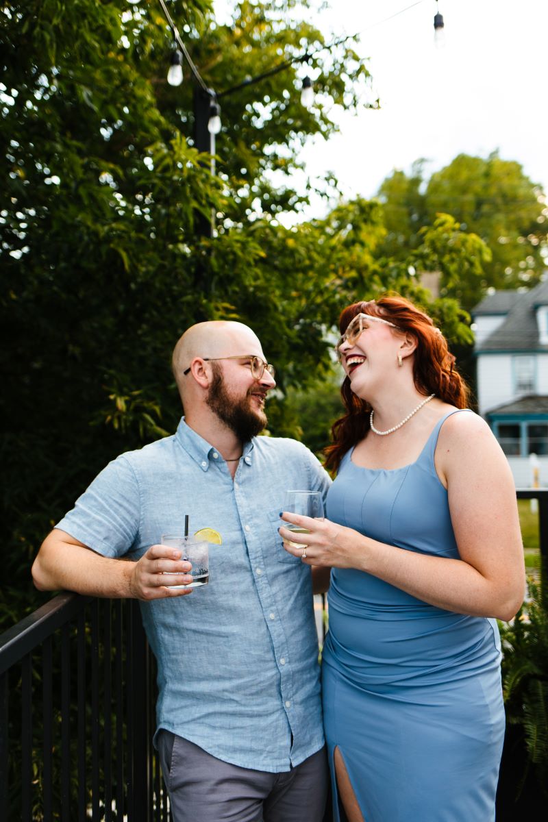 A man in a blue shirt holding a drink in his hand smiling at a woman in a blue dress who is laughing and has red hair and glassess 