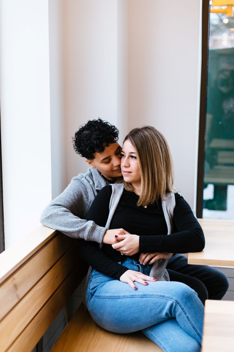 A couple sitting on a wodden bench inside of a coffee shop one is looking ou the window and the other is smiling at their partner 