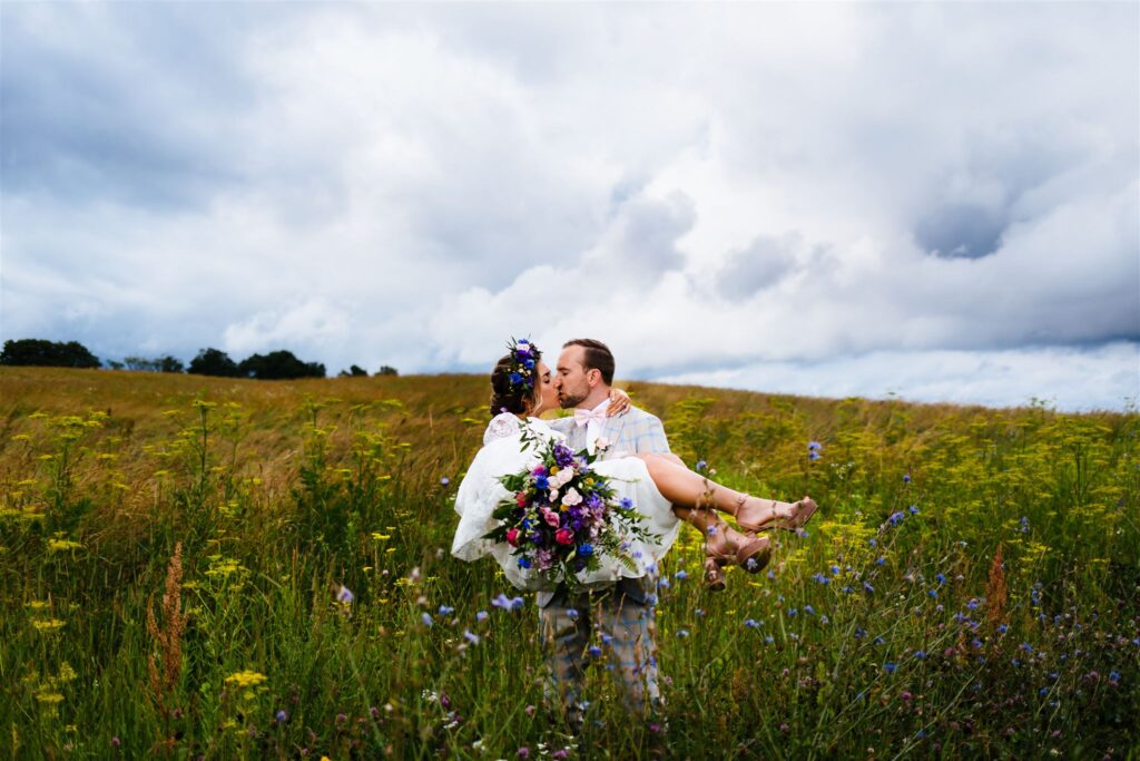 A groom holds his bride who wears a flower crown in a field of wild flowers. They were married at Springside Farm in Fabius NY.