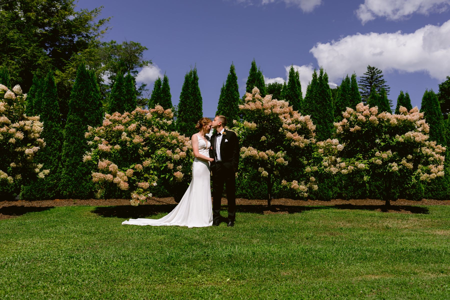 A woman in a white wedding dress is leaning in towards her partner who is wearing a black tuxedo and kissing her forehead behind them are bushes with flowers and green pines and they are standing on grass together