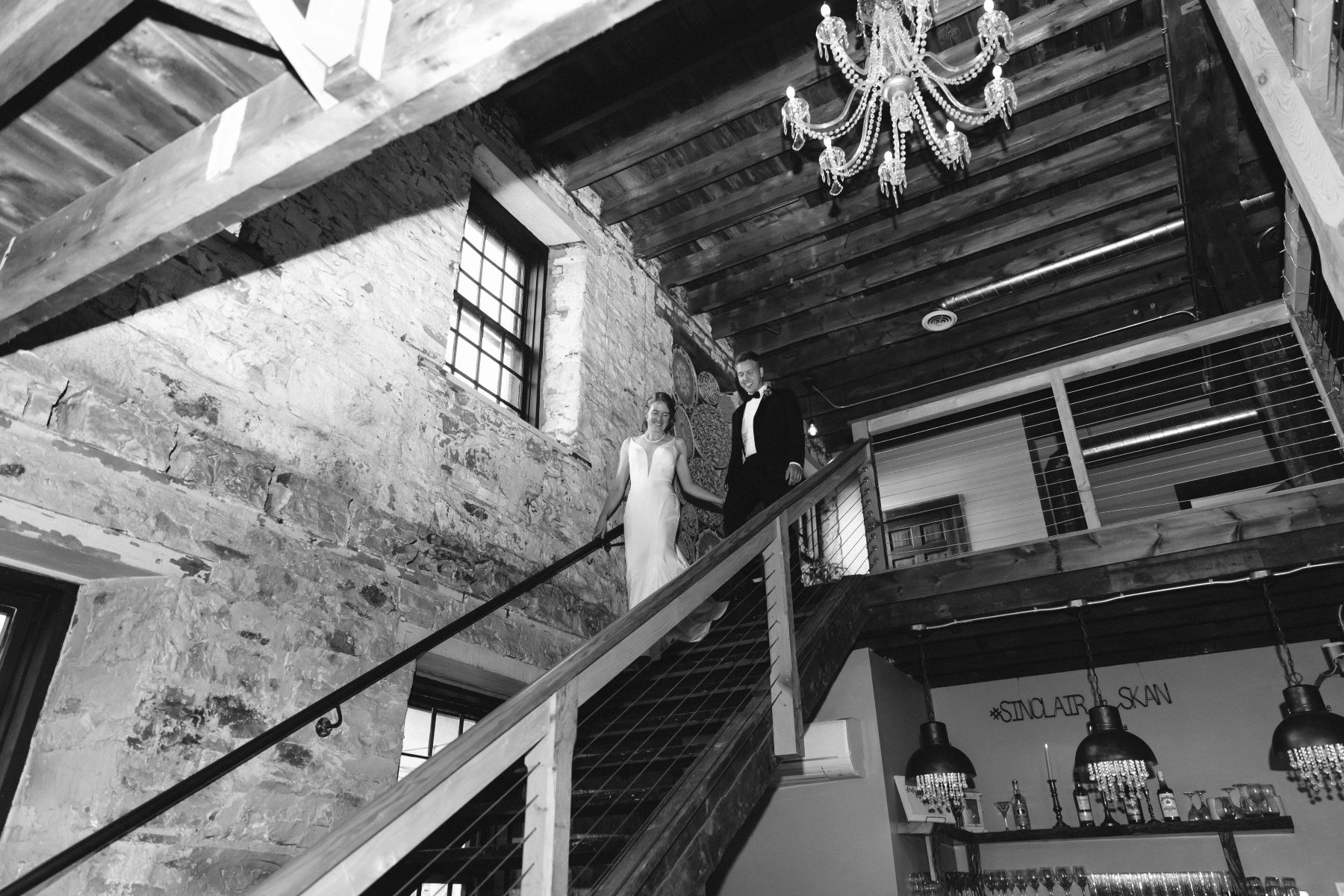 A man in a black suit and bow tie holding hands and walking down the stairs with his wife who is wearing a white wedding dress and a pearl necklace above them are exposed beams and a chandelier and the building is made of stone walls 