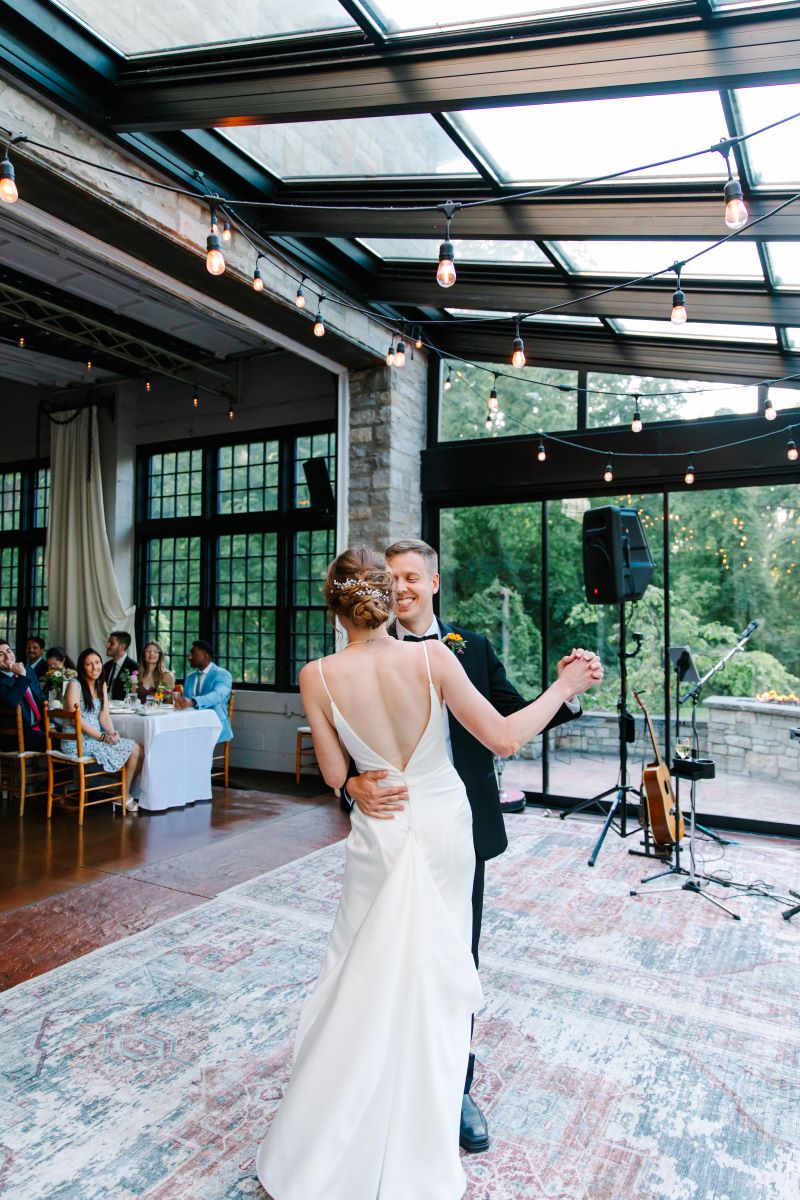 A woman in a white dress is dancing with her husband who is wearing a black tuxedo while their wedding guests watch