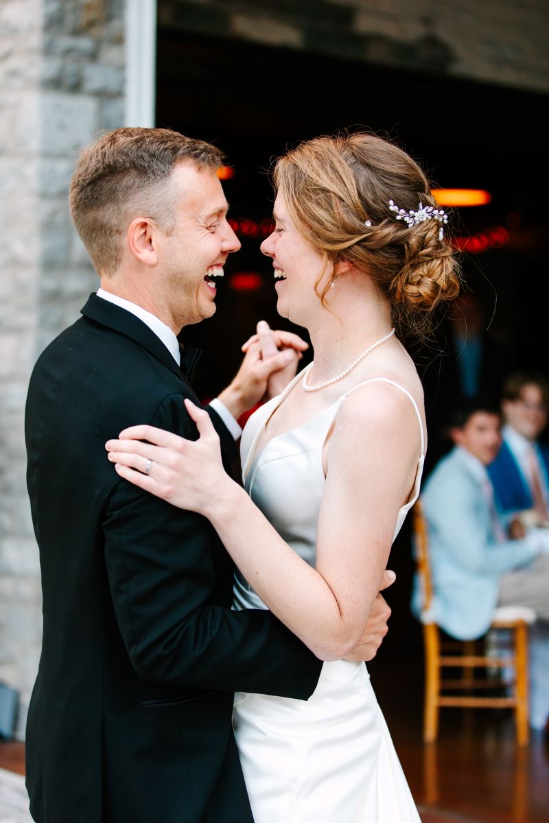 A woman in a white dress with a white pearl necklace is dancing and laughing with her husband who is wearing a black tuxedo while their wedding guests watch