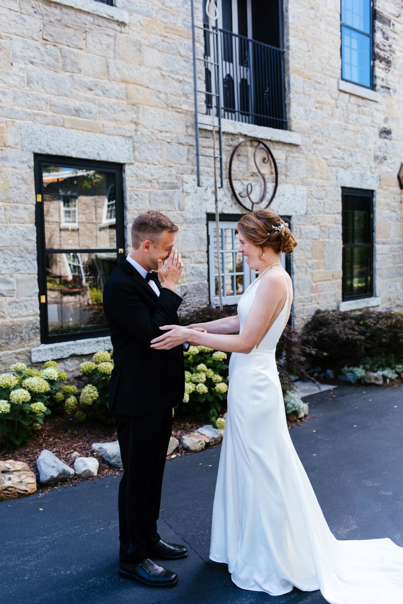 A woman is a whitle wedding dress is touching the elbows of her husband and smiling while her husband who is wearing a black tuxedo is wiping away tears from his eyes 